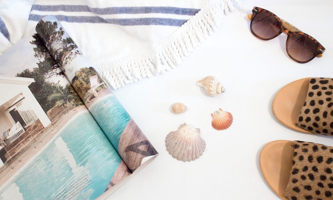opened magazine beside seashells, slide sandals, sunglasses and towel on white surface