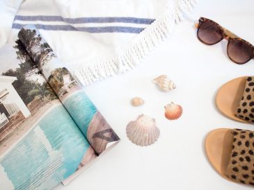 opened magazine beside seashells, slide sandals, sunglasses and towel on white surface
